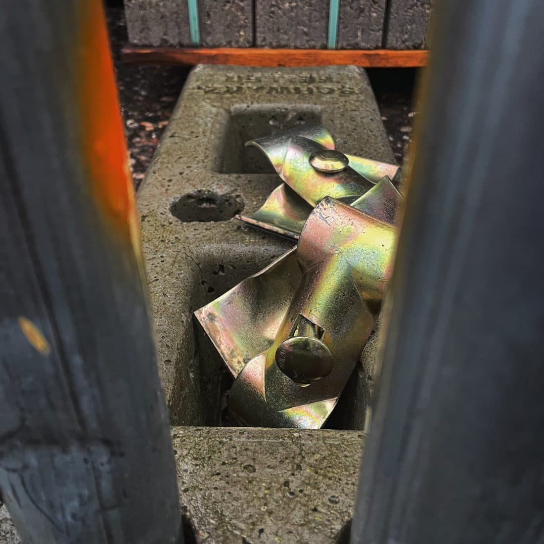 Schillernde, gelbgrün glänzende Metallteile liegen in der Aussparung eines Betonblocks, gesehen durch einen schmalen Spalt zwischen zwei dunklen Metallpfosten