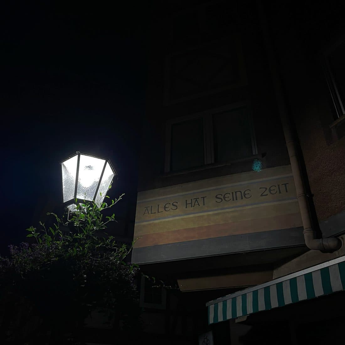 Eine leuchtende Straßenlaterne in der Nacht, daneben eine dunkle Hausfassade mit der aufgemalten Inschrift „Alles hat seine Zeit“ und einer grün-weiß gestreiften Markise