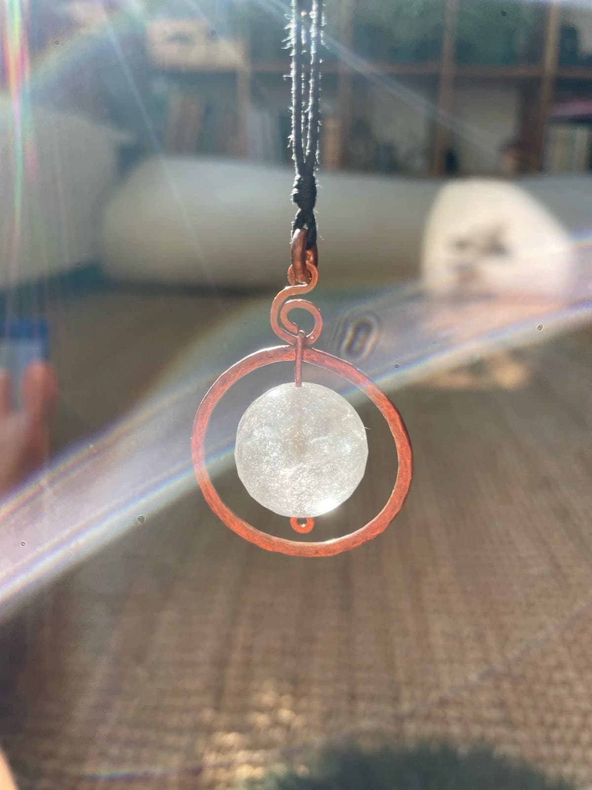 Facettierte, milchig durchscheinende Bergkristallkugel, gehalten in einem gehämmerten Kupferring, darüber eine Kupferspirale, an schwarzem Band; im Gegenlicht mit Regenbogenreflexen