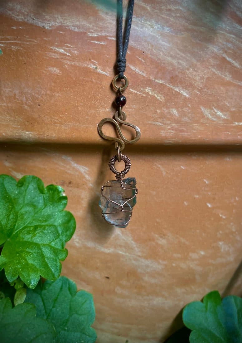 Eine bläulich schimmernde Glasscherbe, mit Kupferdraht umwickelt, darüber ein flachgehämmertes geschwungenes Kupferelement und eine dunkelrote Granatperle, an dunkler Schnur vor Terracotta