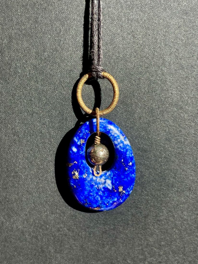 Ein tiefblauer Lapislazuli als ovale Scheibe mit tropfenförmiger Öffnung, darin eine kleine goldglänzende Pyritkugel an Messingdraht, darüber eine gewickelte Messingöse an schwarzer Schnur vor grauem Grund