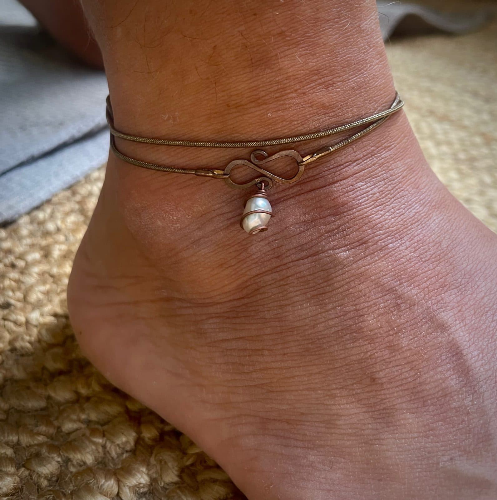 Fußkette, zweifach um einen Knöchel gelegt, mit einem gehämmerten Unendlichkeitszeichen und einer weißen Perle, die in Kupferdraht gewickelt ist