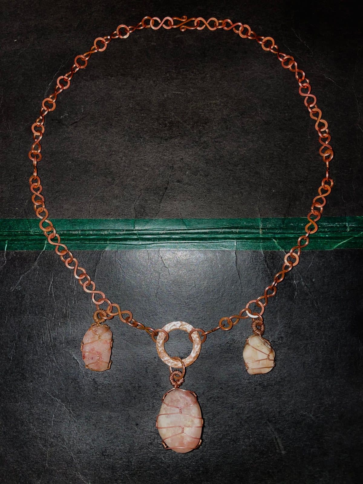 Eine Kette aus Unendlichkeitszeichen und kleinen Ringen aus Kupfer, im Kreis auf dunklem Untergrund ausgelegt; unten hängen drei rosafarbene Steine in Kupferdraht, der größte an einem hellen Ring in der Mitte