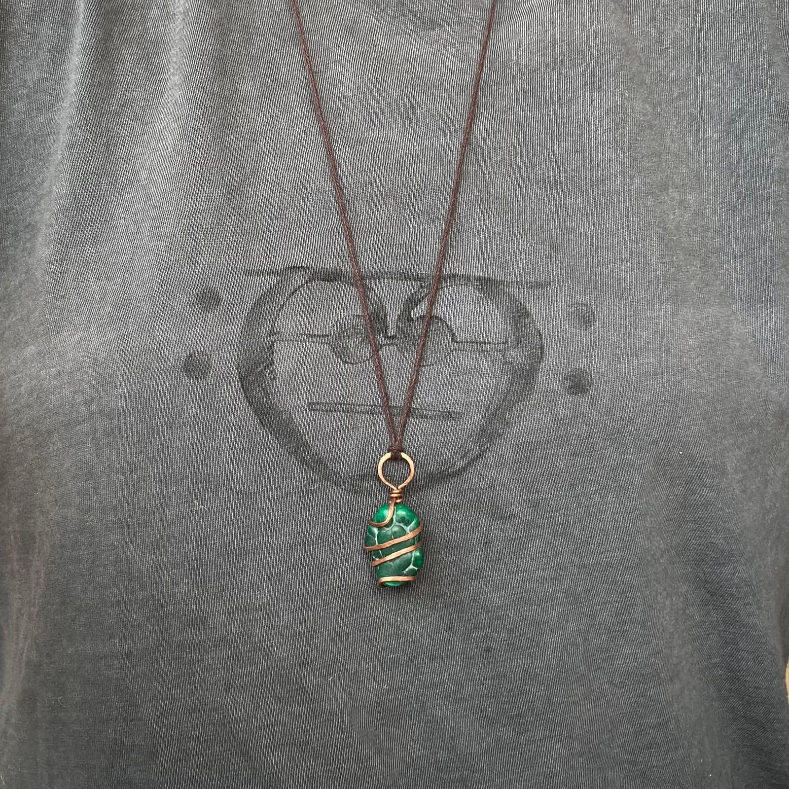 Ein grüner Malachit, spiralförmig mit Kupferdraht umwickelt, an einer braunen Baumwollkordel, getragen über einem grauen T-Shirt mit aufgedrucktem Motiv