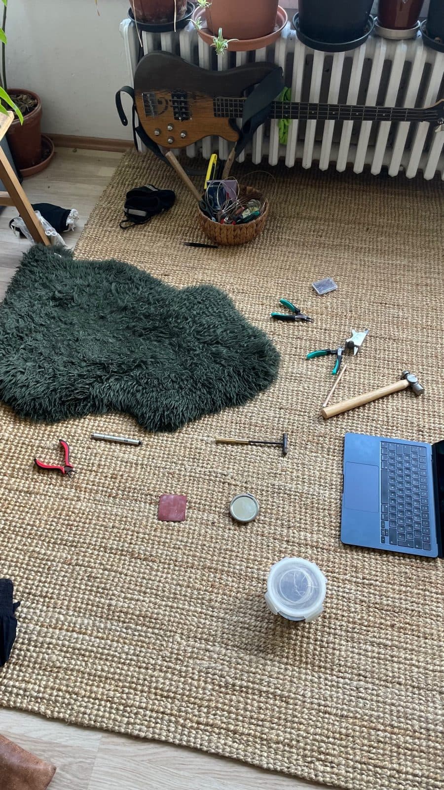 Der Arbeitsplatz auf dem Boden: Juteteppich mit grünem Schaffell, verstreute Zangen, zwei Hämmer, ein kleiner Amboss, ein Werkzeugkorb und ein Laptop; an der Heizung dahinter lehnt ein E-Bass